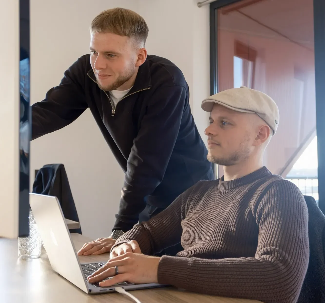 Twee mannen in modern kantoor, een staand bij bureau en een zittend achter laptop met smartphone op tafel.