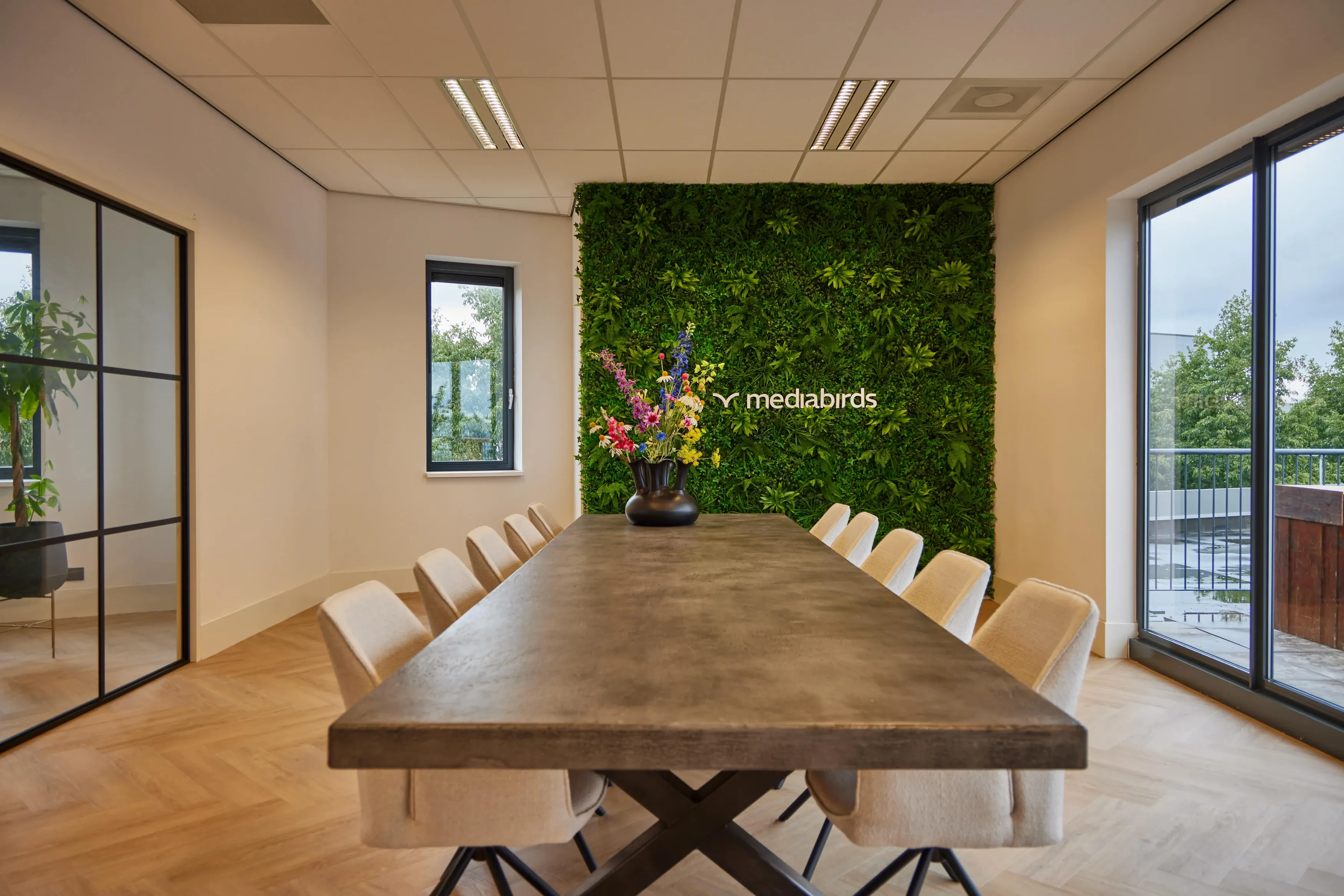 Moderne vergaderruimte met lange industriële eettafel, lichtgekleurde stoelen en groene wand met mediabirds-logo
