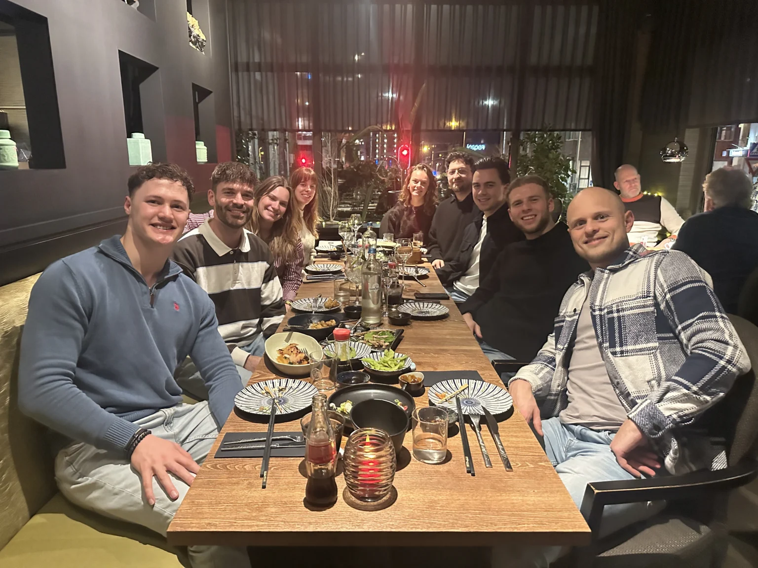 Tien personen zitten lachend en pratend rond een eettafel in een modern restaurant met donkere muren.
