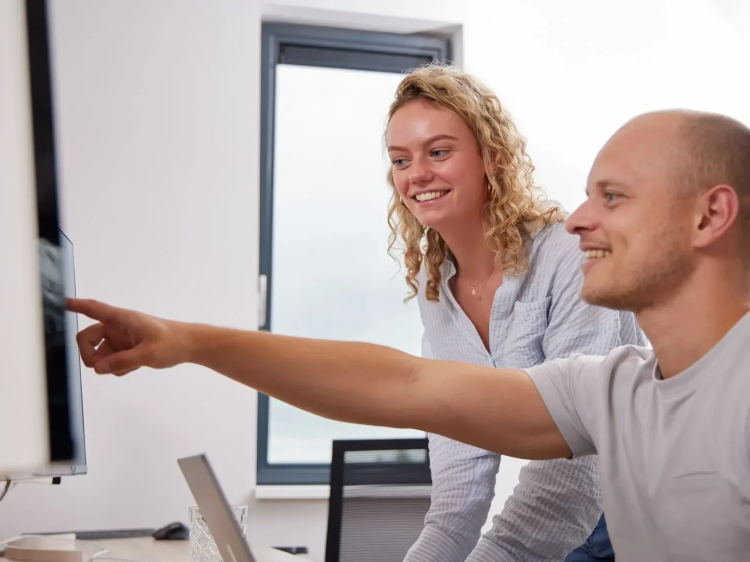 Man in lichtgrijs t-shirt en vrouw met krullend haar en gestreepte blouse werken samen aan een bureau