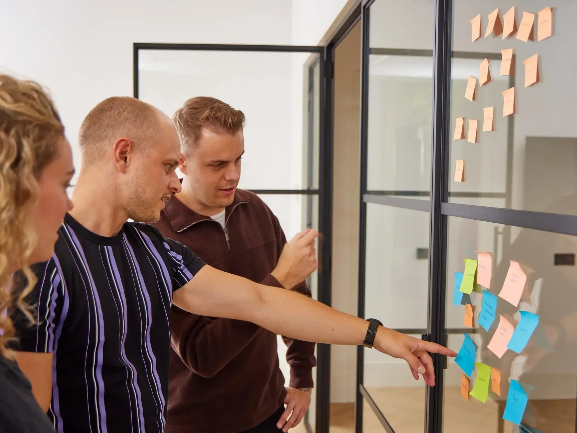Drie mensen bij een glazen wand met kleurrijke post-its in een lichte, moderne werkomgeving.