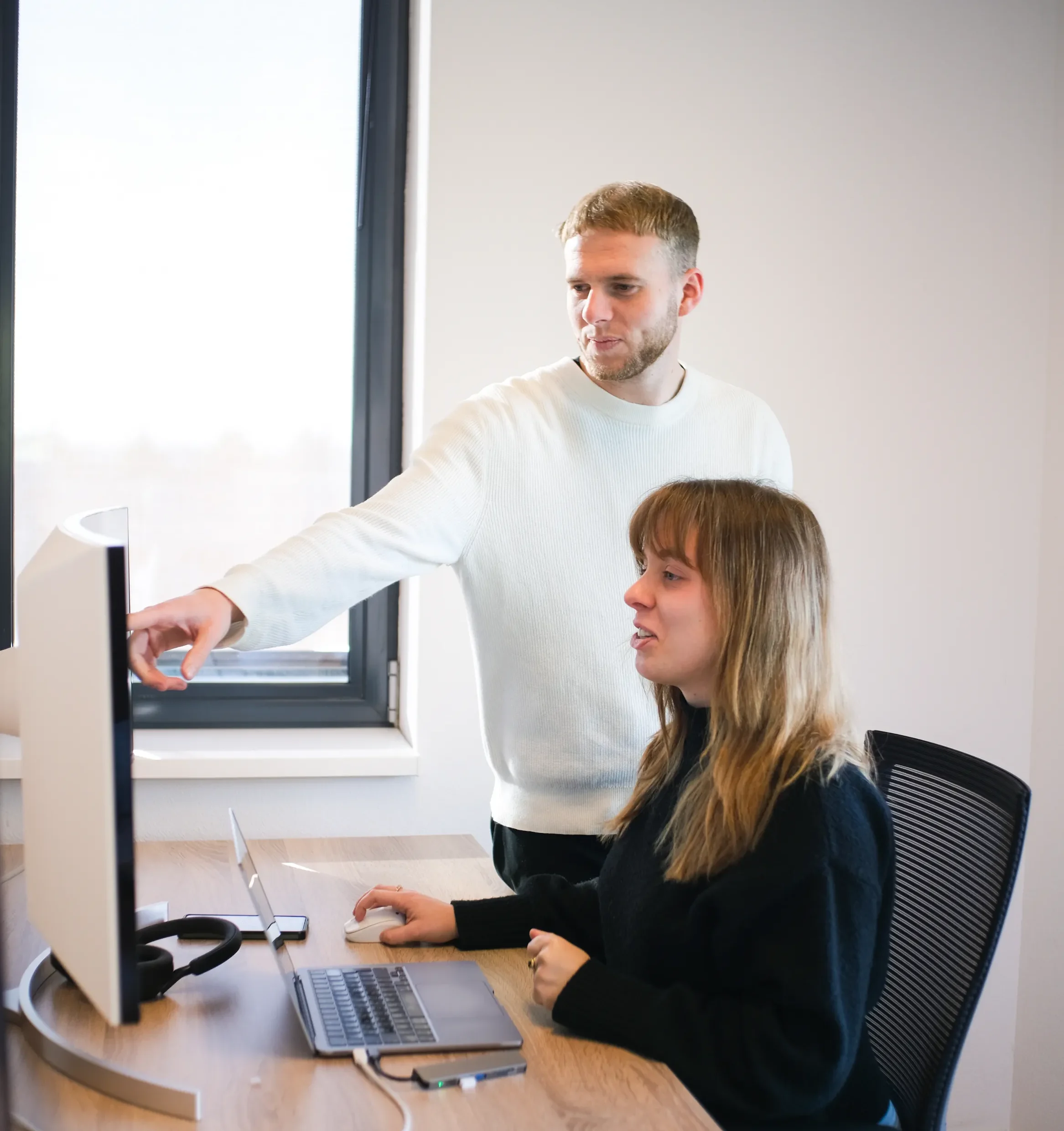 Een man in witte trui wijst naar laptopscherm terwijl een vrouw in zwarte trui ernaar kijkt in kantoor