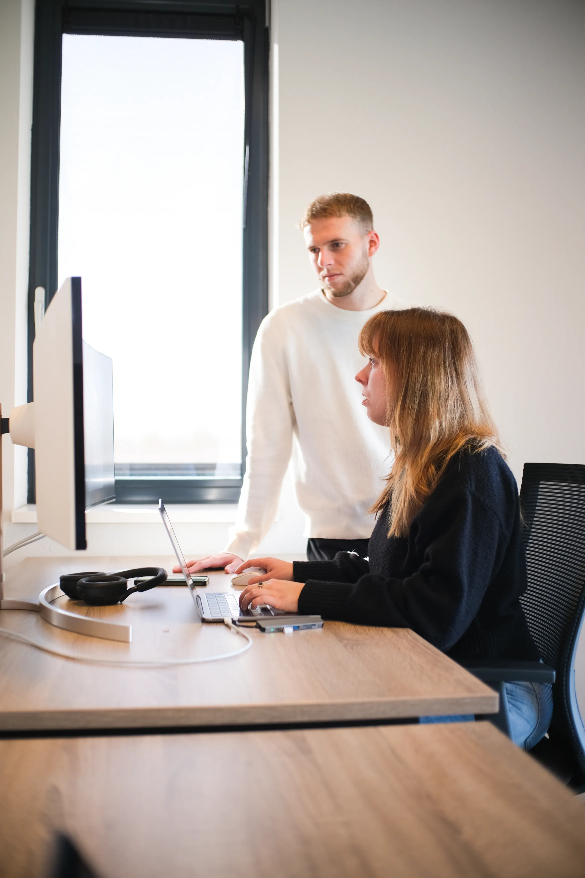 Lichte moderne kantoorruimte met twee mensen bij een houten bureau, vrouw werkt op laptop, man kijkt mee.