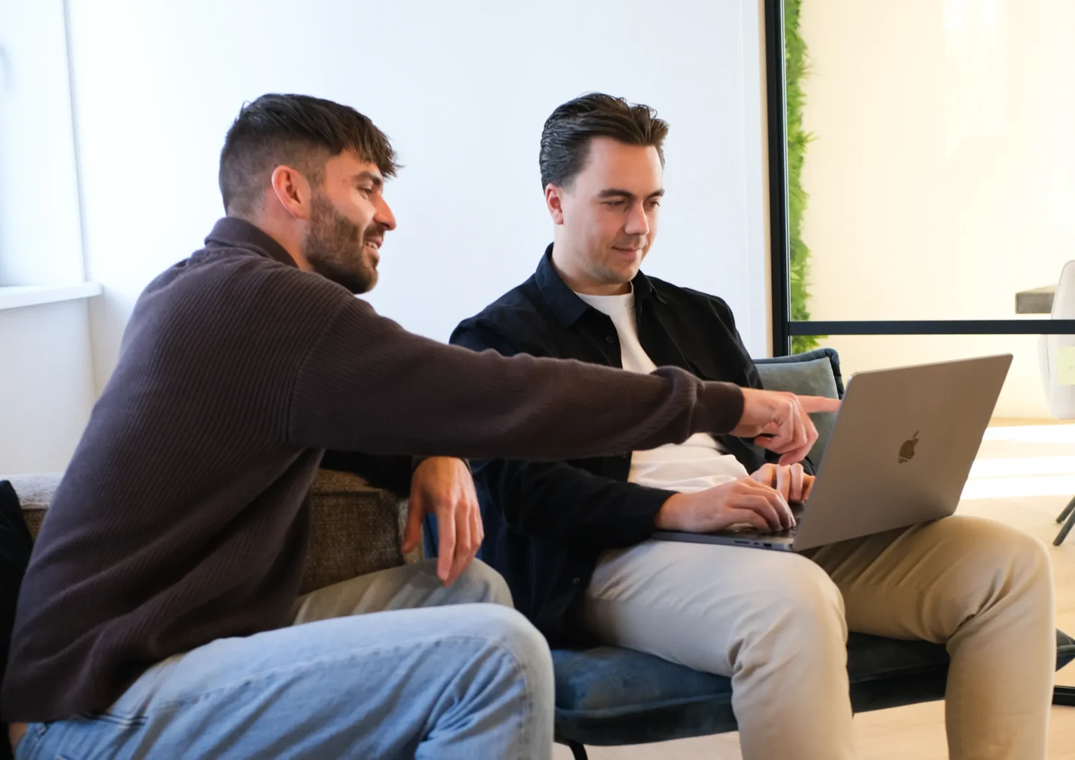 Twee jonge mannen in casual kleding werken samen op een laptop in een lichte, moderne ruimte met grote ramen.