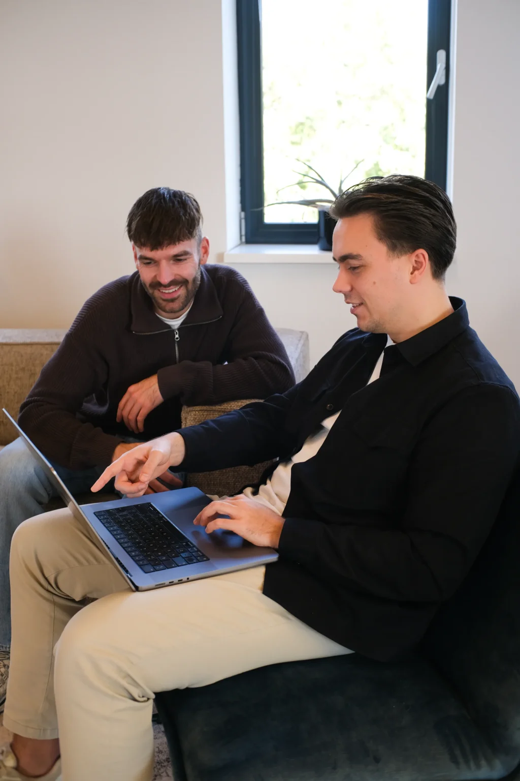 Twee mannen zitten op een bank, één typt op laptop, de ander wijst lachend naar het scherm