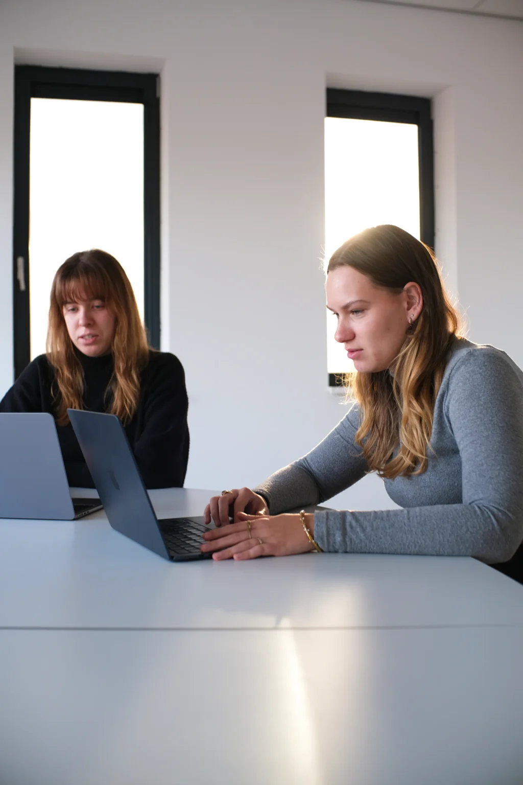 Twee vrouwen zitten aan een tafel in een lichte, moderne ruimte, beiden met laptops.
