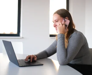 Vrouw zit geconcentreerd aan haar bureau met laptop en telefoon in een lichte, moderne ruimte.