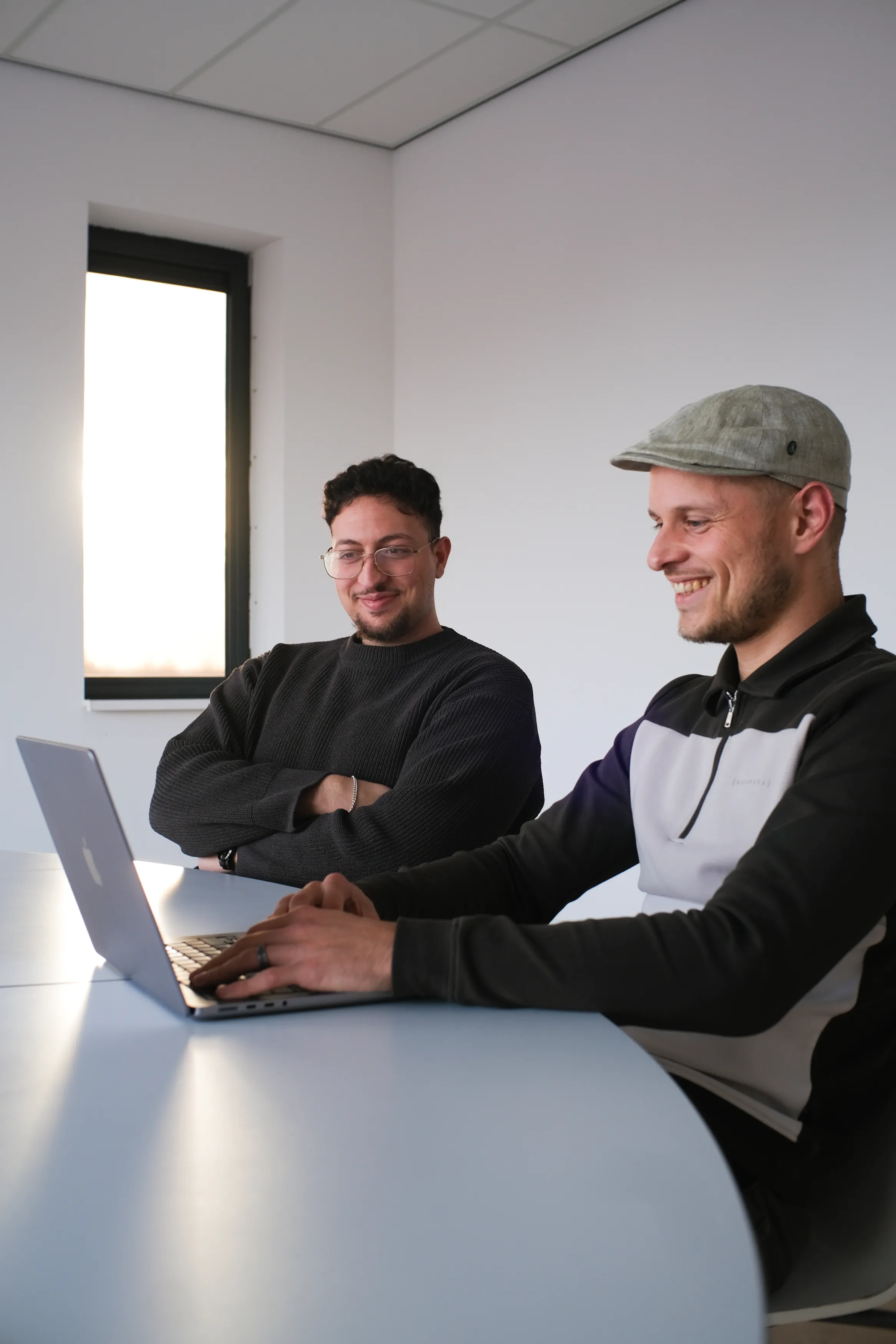 Twee mannen met glimlach zittend aan tafel met laptop in moderne lichte kantoorruimte