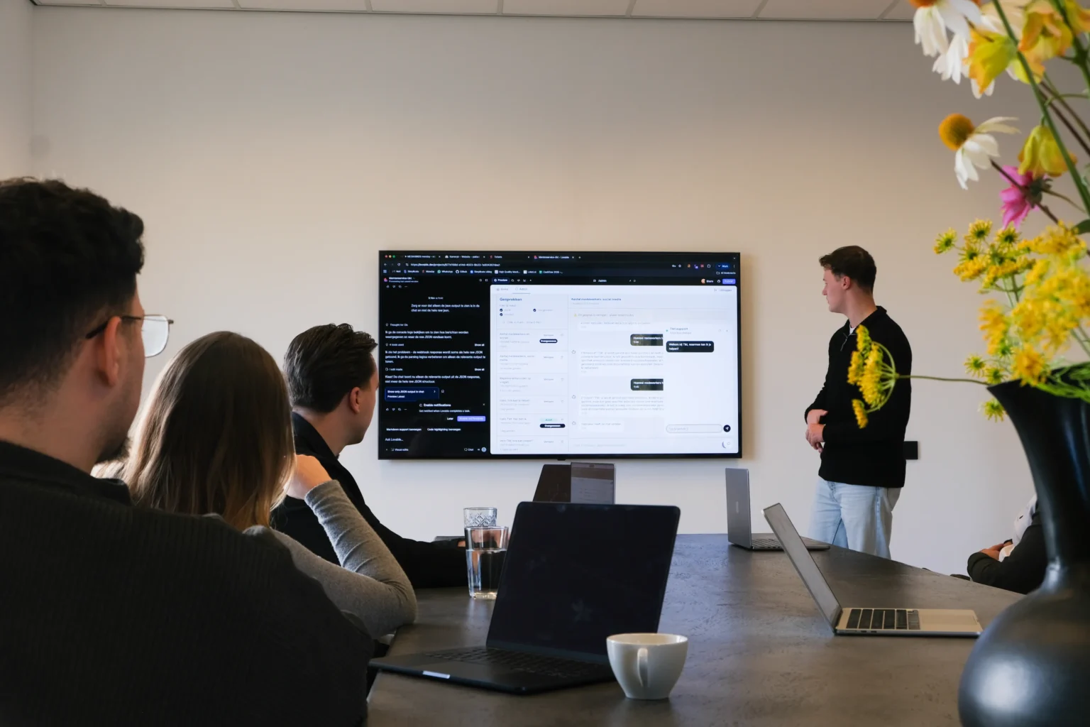 Vier mensen in een moderne vergaderruimte kijken naar een groot scherm met tekst en chats.