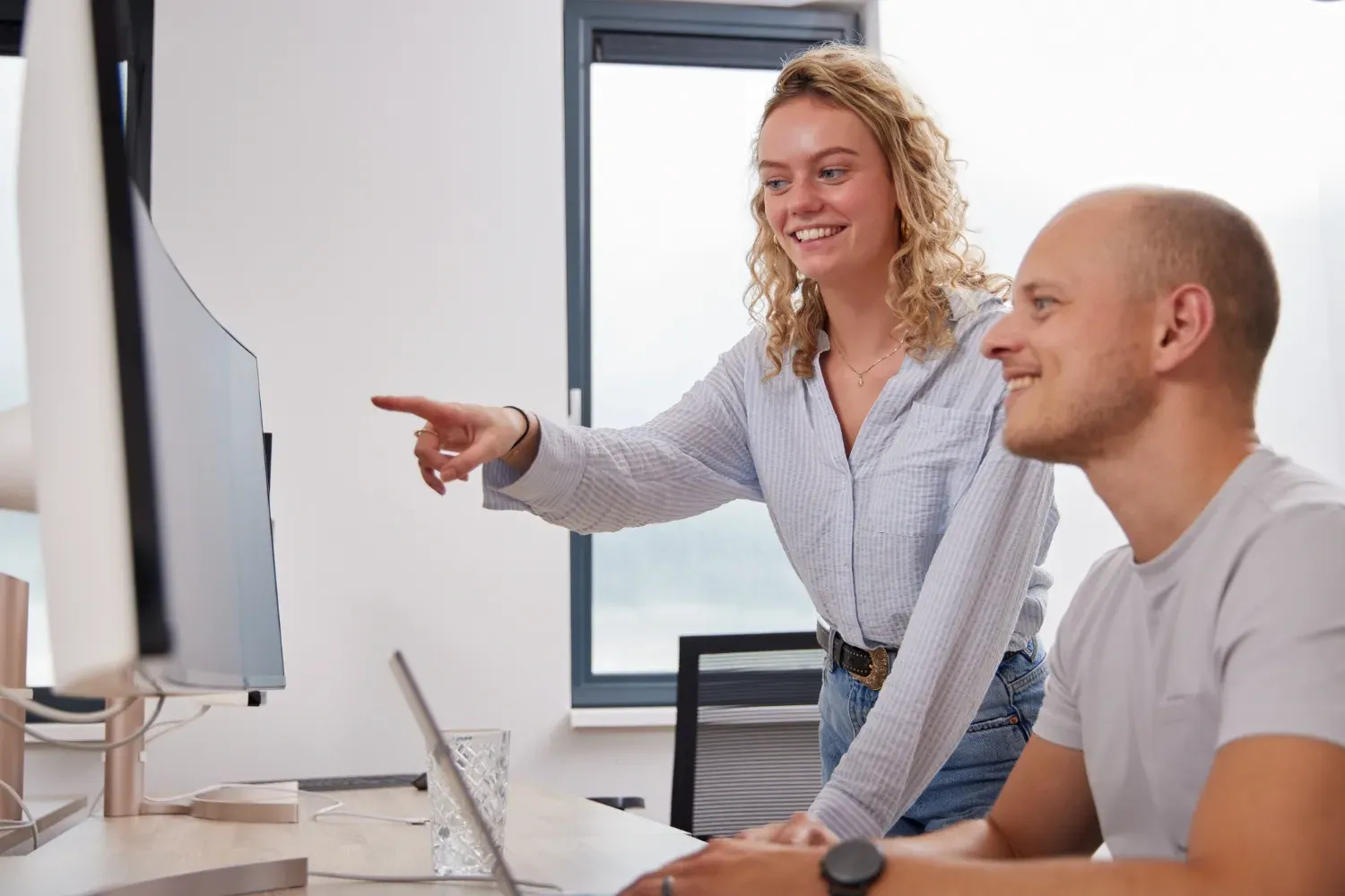 Twee personen in kantoor: vrouw wijst naar computerscherm, man glimlacht naast bureau met laptop