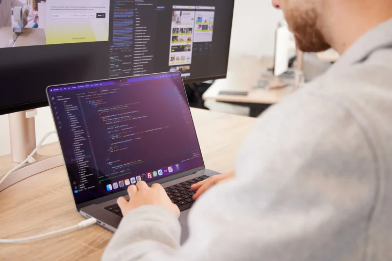 Persoon werkt geconcentreerd aan laptop met code op scherm, extern beeldscherm toont webpagina's