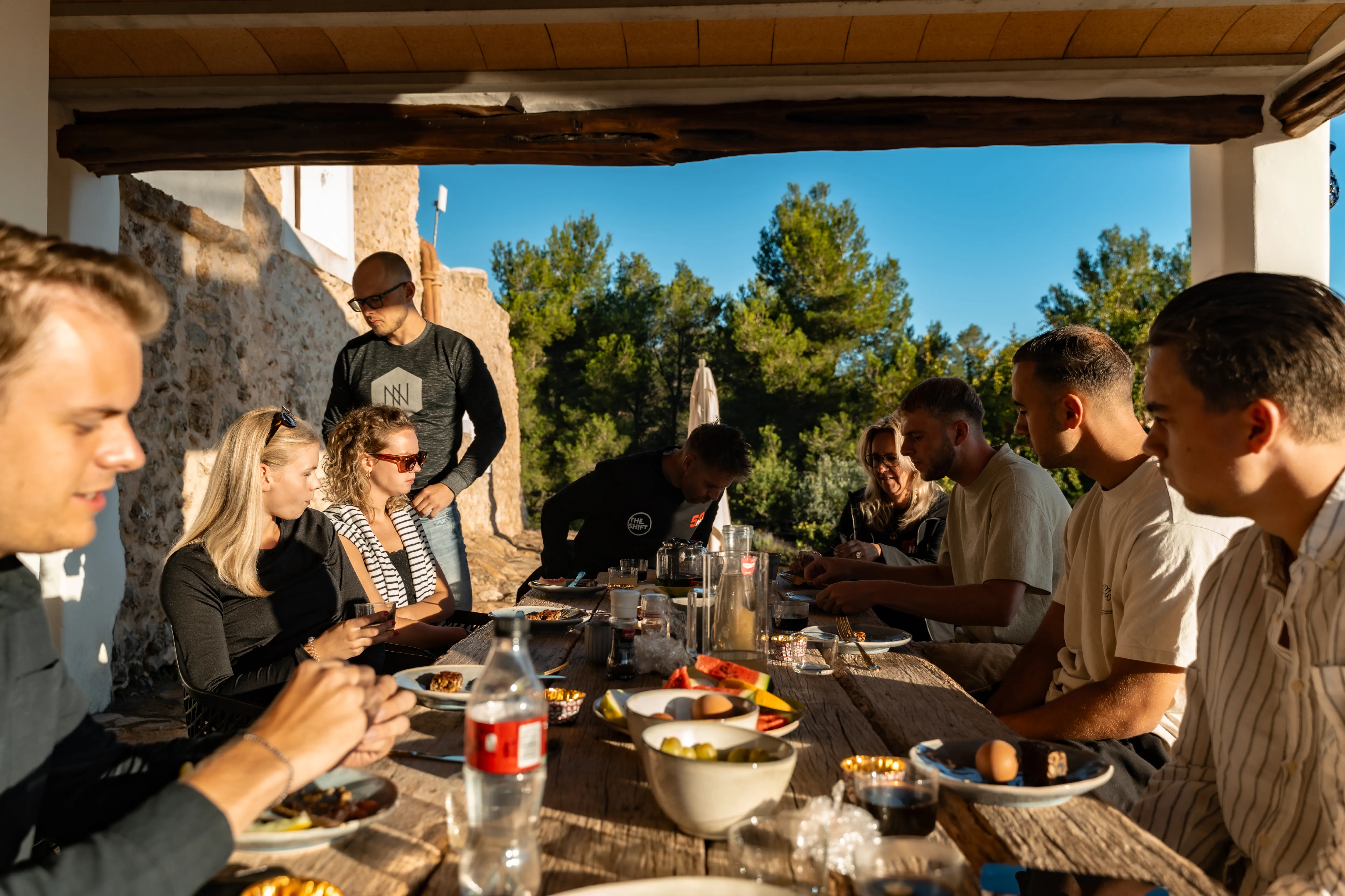 Groep mensen aan lange houten tafel op terras met eten en glaswerk in groene omgeving.