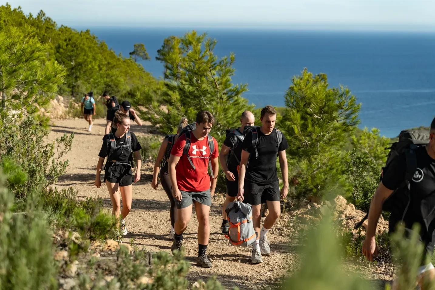 Een groep mensen wandelt op een onverhard pad omgeven door groene bomen met uitzicht op de zee.