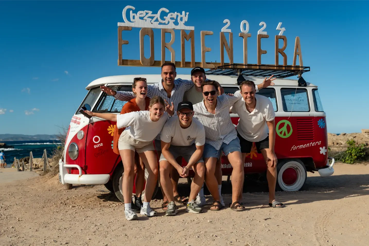 Zeven mensen poseren lachend voor een rode Volkswagen bus met tekst Formentera 2024