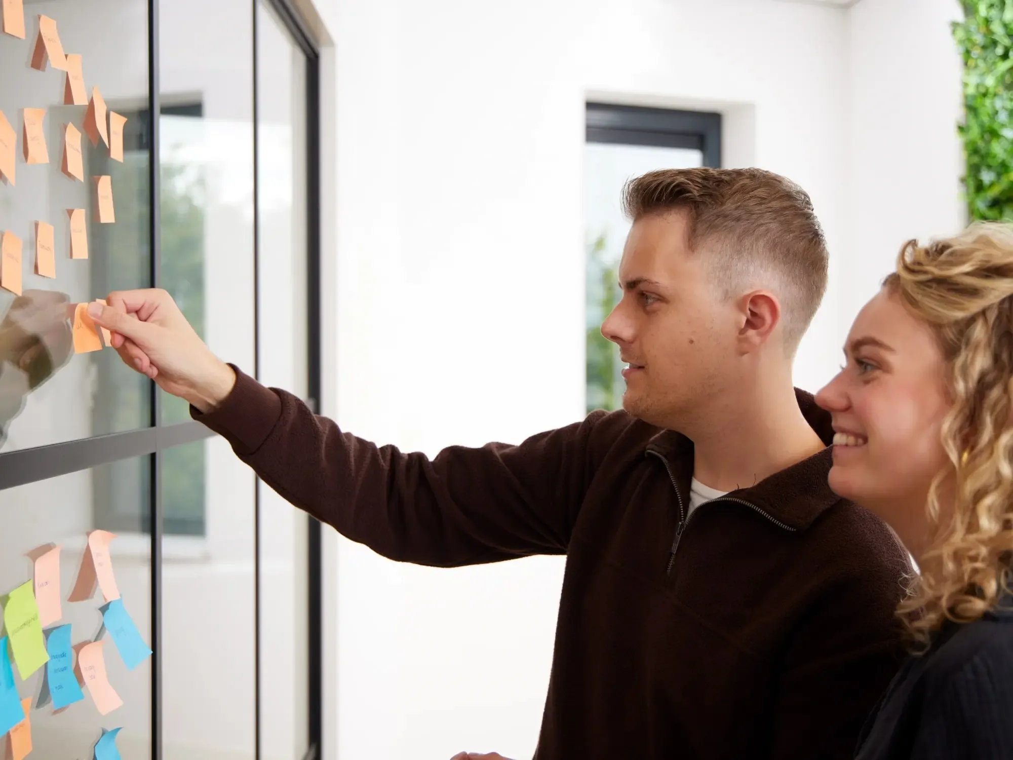 Man hangt gele post-it op glazen wand met gekleurde post-its terwijl vrouw glimlacht naast hem