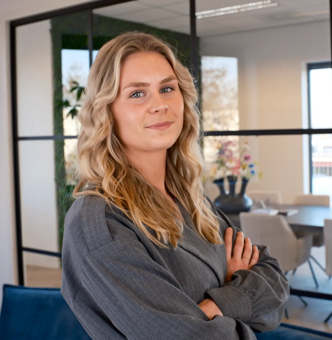Vrouw met lang golvend haar en grijze blazer kijkt zelfverzekerd met gekruiste armen in modern kantoor