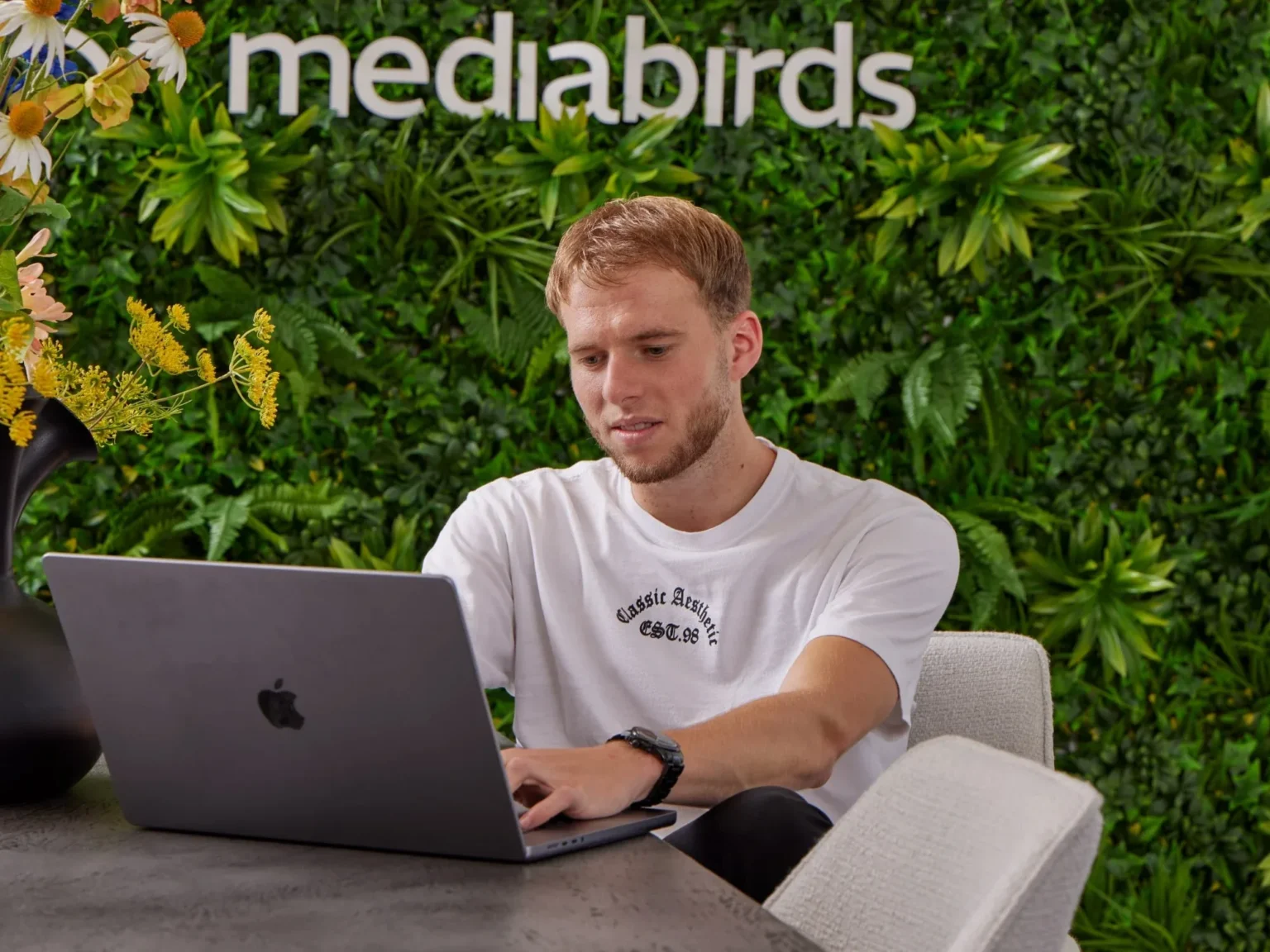 Jongeman in wit t-shirt werkt geconcentreerd achter laptop aan tafel met zwarte vaas en planten.