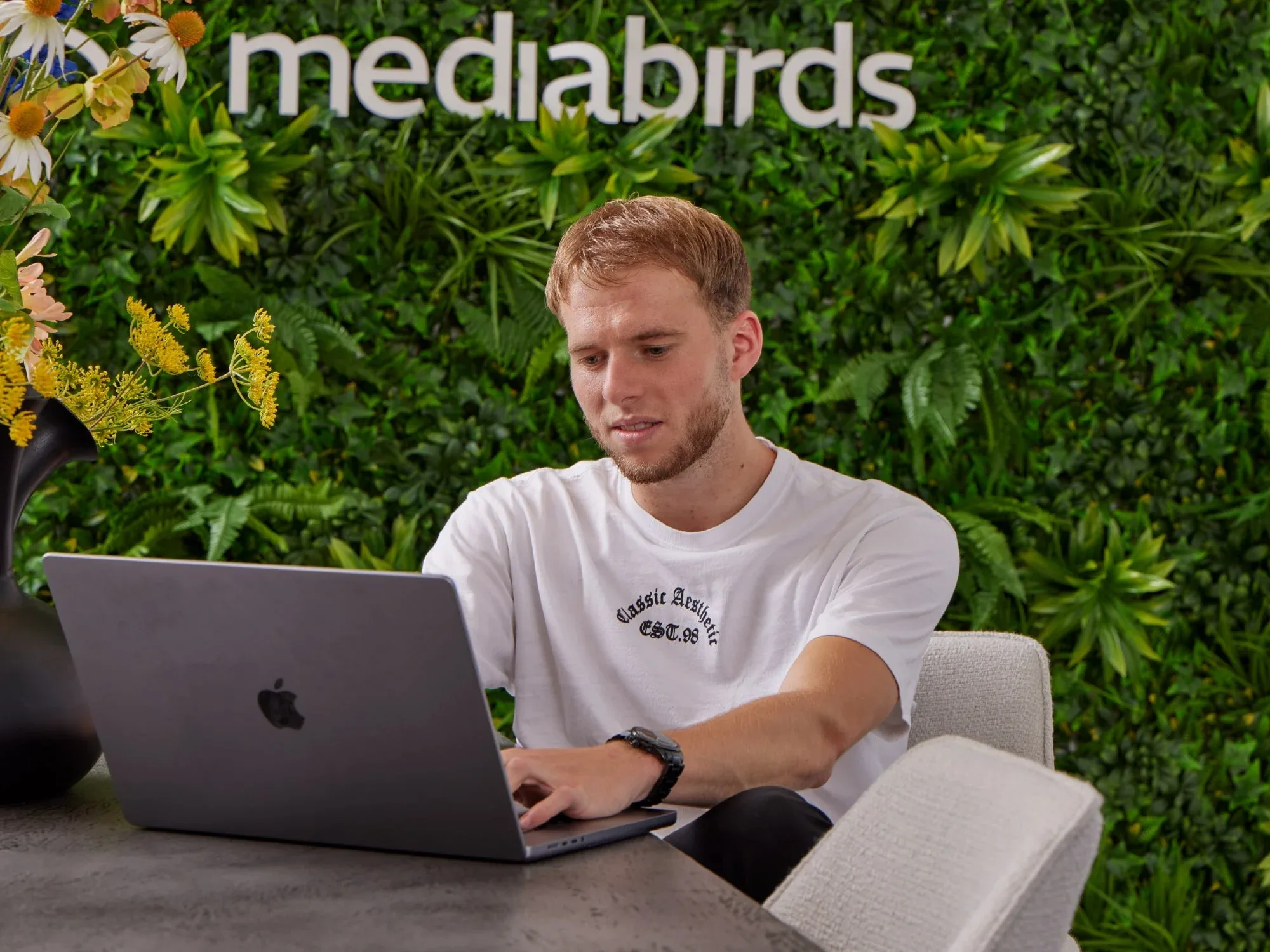 Jongeman in wit t-shirt werkt geconcentreerd achter laptop aan tafel met zwarte vaas en planten.