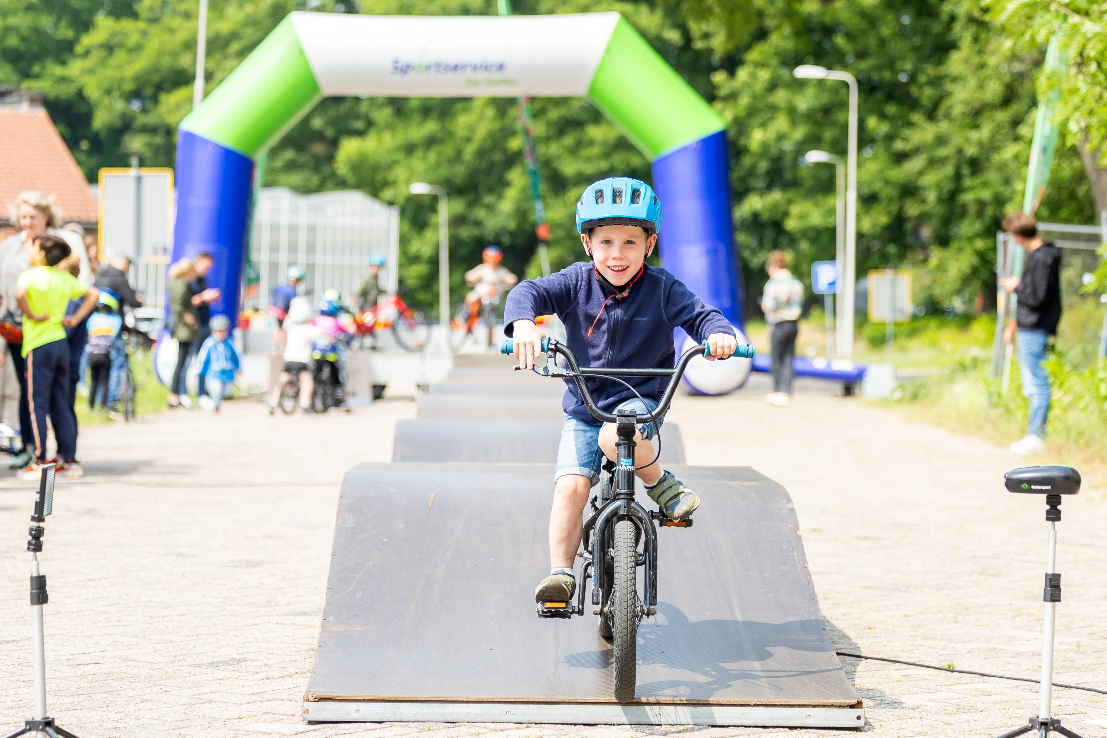 Jongen met blauwe fietshelm rijdt glimlachend op fiets over helling bij sportevenement met kleurrijke boog