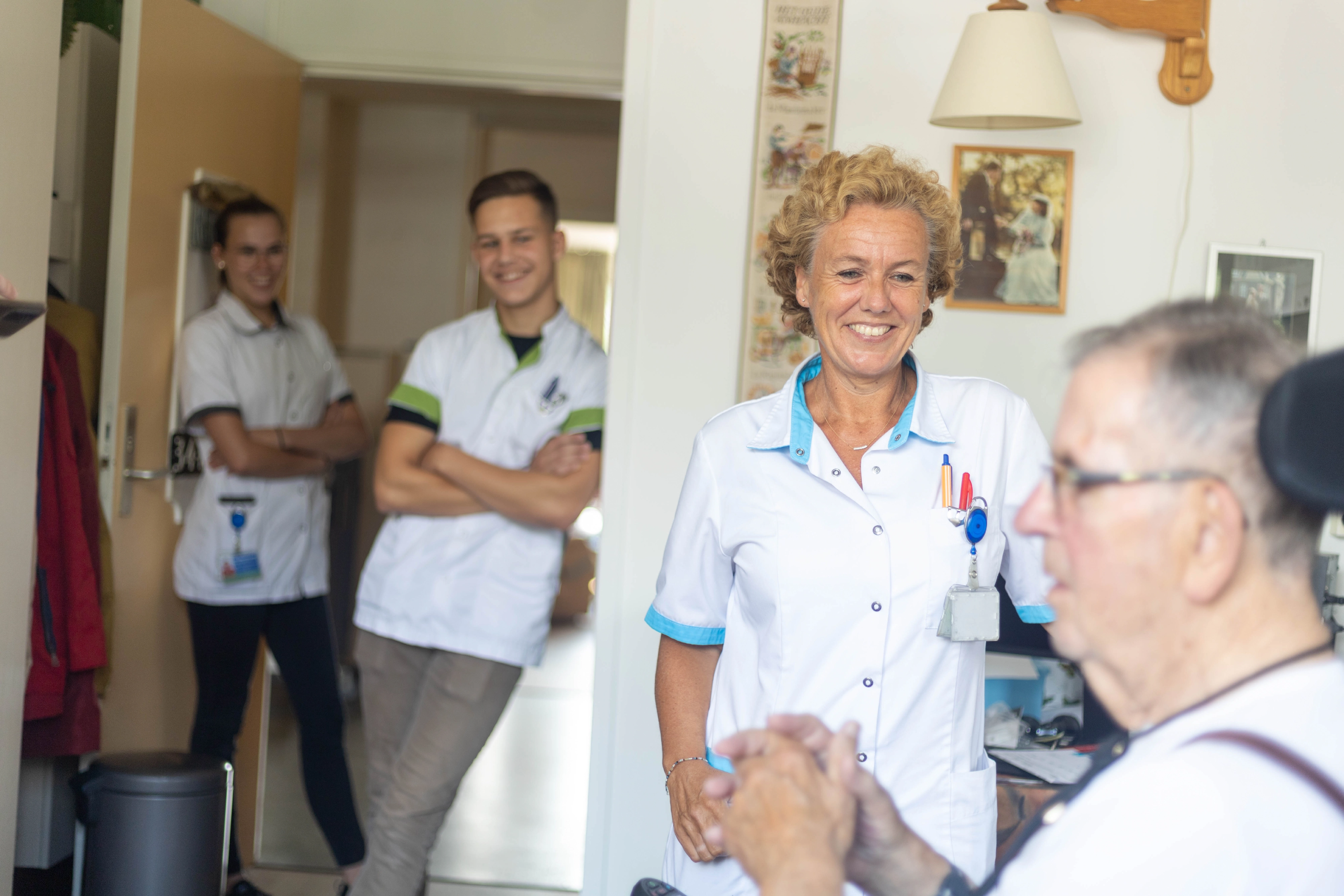 Vier mensen in een huiselijke ruimte, waaronder een oudere man in een rolstoel en drie verpleegkundigen die samen praten
