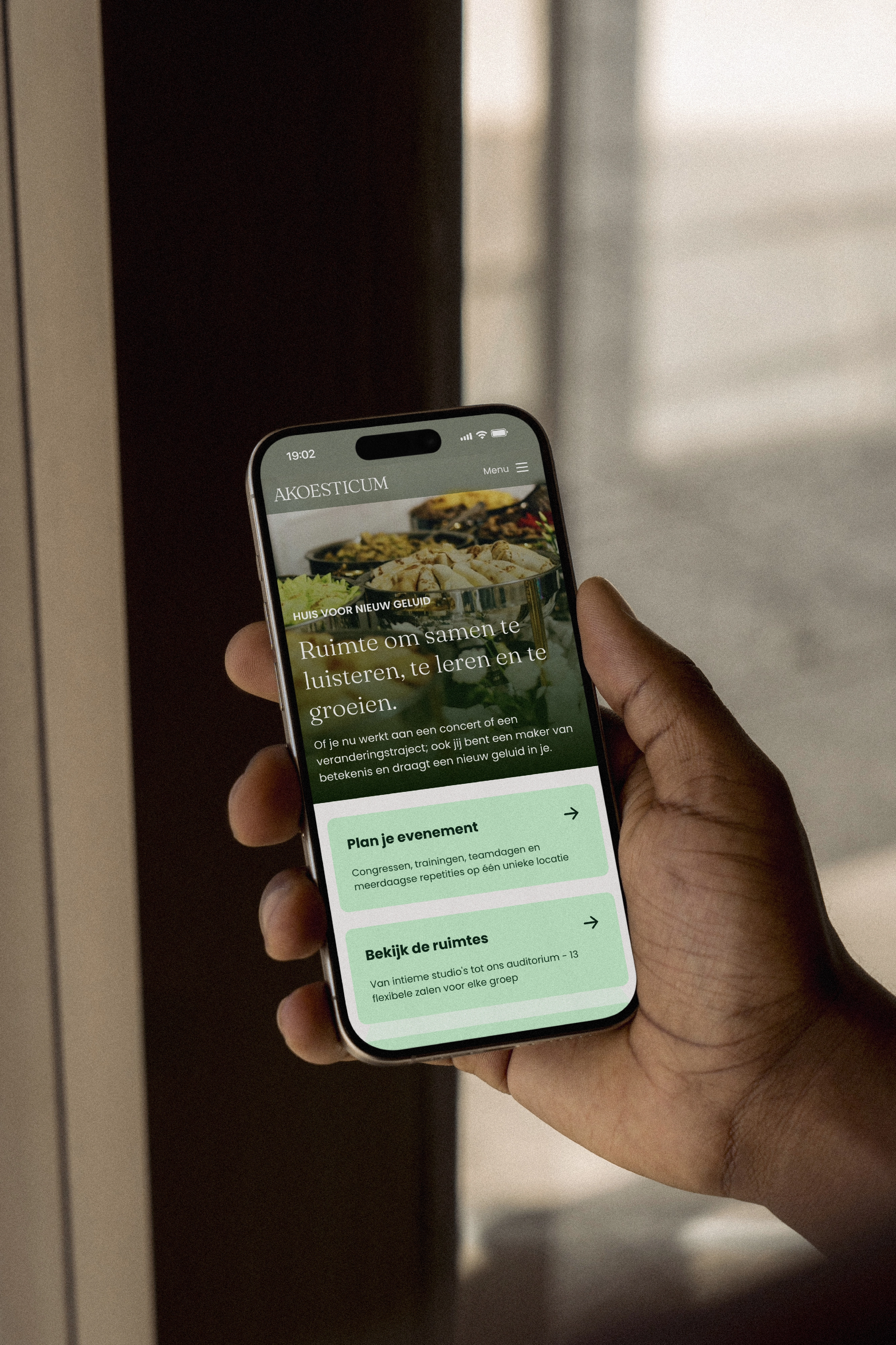 Hand houdt een smartphone met daarop een website 'AKOESTICUM' en opties voor evenementen en ruimtes.
