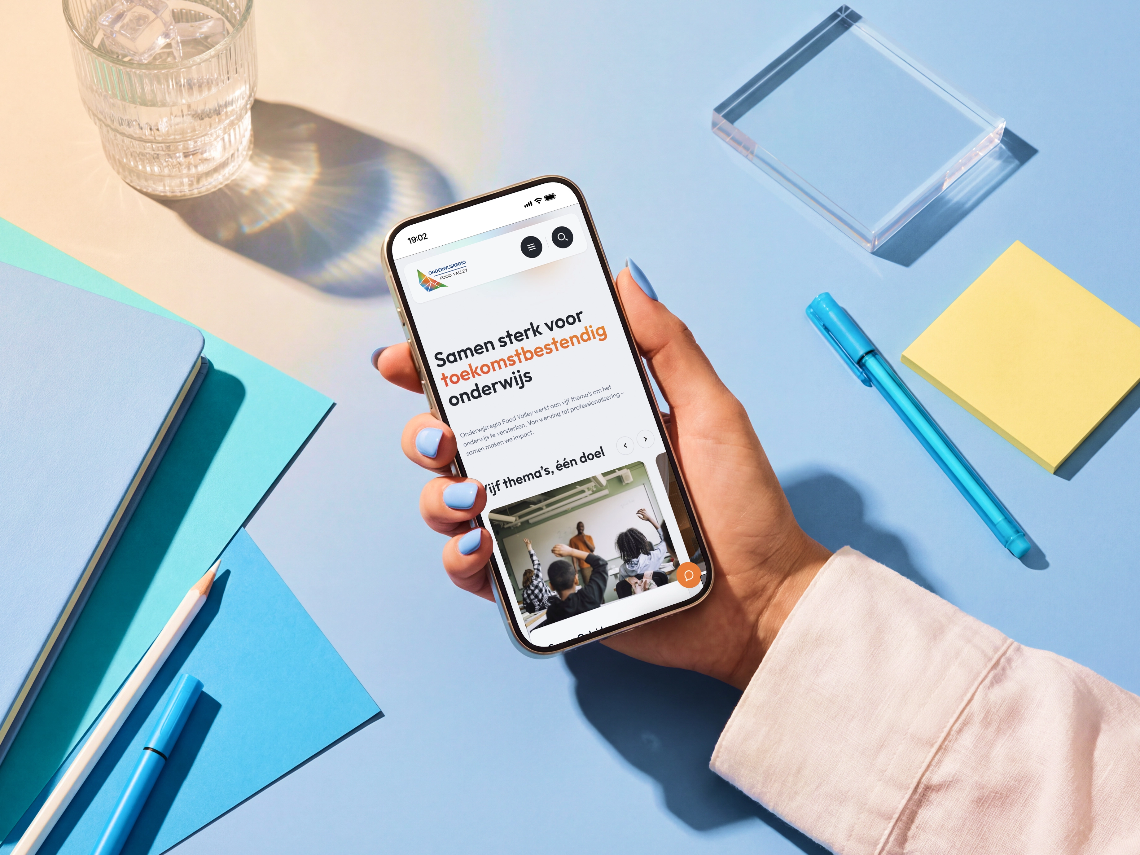 Hand houdt smartphone met tekst 'Samen sterk voor toekomstbestendig onderwijs' en kantoorartikelen